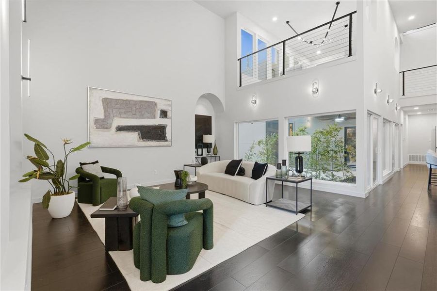 Double-height living space featuring dark wood-finish flooring, a contemporary staircase with cable railing, large windows with exterior foliage, and recessed lighting