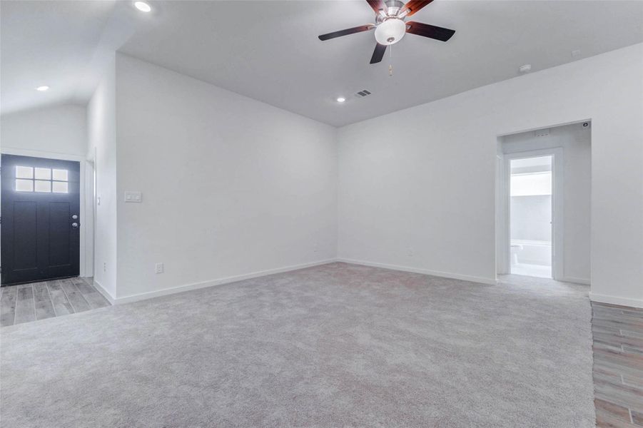 Large family room view with plush carpet, high ceilings, ceiling fan, recessed lighting, and sightlines to the entry and adjoining spaces. The neutral walls and open layout create a flexible blank canvas for comfortable seating, media, décor, and personal style.