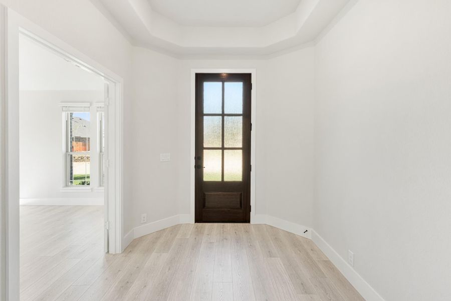 Empty room with tray ceiling, light wood floors, and dark wood door with glass panels leading outside.