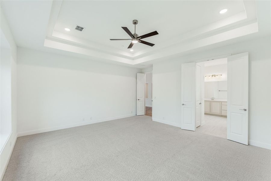 Empty bedroom with tray ceiling, ceiling fan, carpet flooring, and open doors to bathroom and closet