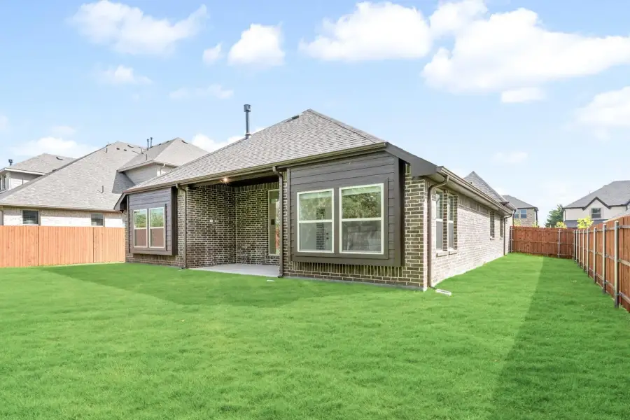 Rear view of single-story brick home with covered patio, large windows, and fenced green backyard