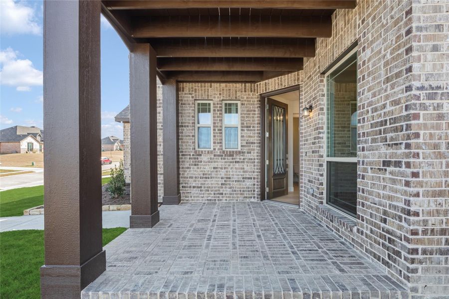 Covered front porch with brick flooring, dark wood beams, and decorative front door on brick exterior home Covered front porch with brick flooring, dark wood beams, and decorative front door on brick exterior home
