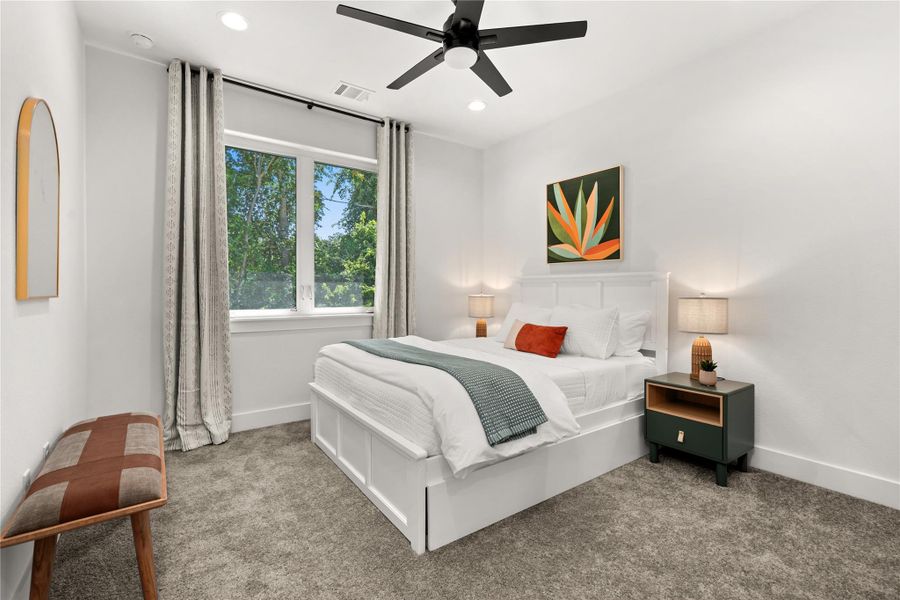 This cozy bedroom features a modern design with ample natural light from a large window. It includes a stylish ceiling fan, and soft carpeting, creating a welcoming and comfortable atmosphere.
