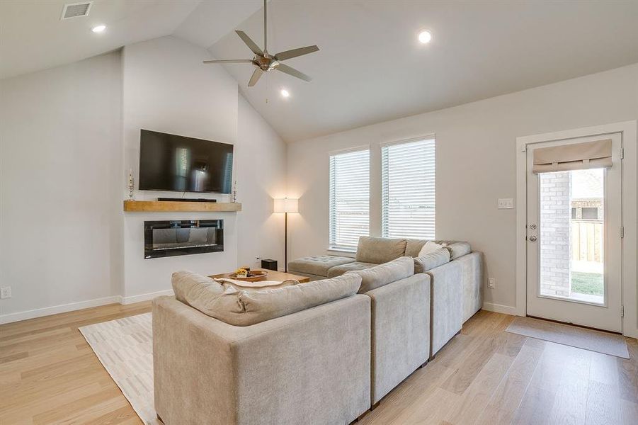 Living room with plenty of natural light, light wood-style flooring, high vaulted ceiling, a ceiling fan, and a glass covered fireplace Living room with plenty of natural light, light wood-style flooring, high vaulted ceiling, a ceiling fan, and a glass covered fireplace