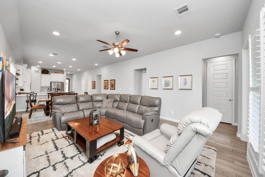 Spacious open-concept living area with modern decor, featuring a comfortable sectional, ceiling fan, and adjacent kitchen. Natural light streams in through large windows, highlighting the neutral tones and sleek finishes.