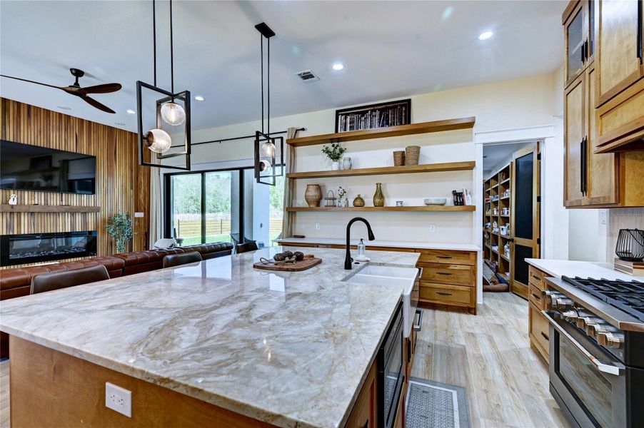 This modern kitchen features a spacious marble island with a sink, stylish pendant lighting, and ample wooden cabinetry. It opens to a cozy living area with a wood-accented wall, fireplace, and large windows, offering a bright and inviting space.