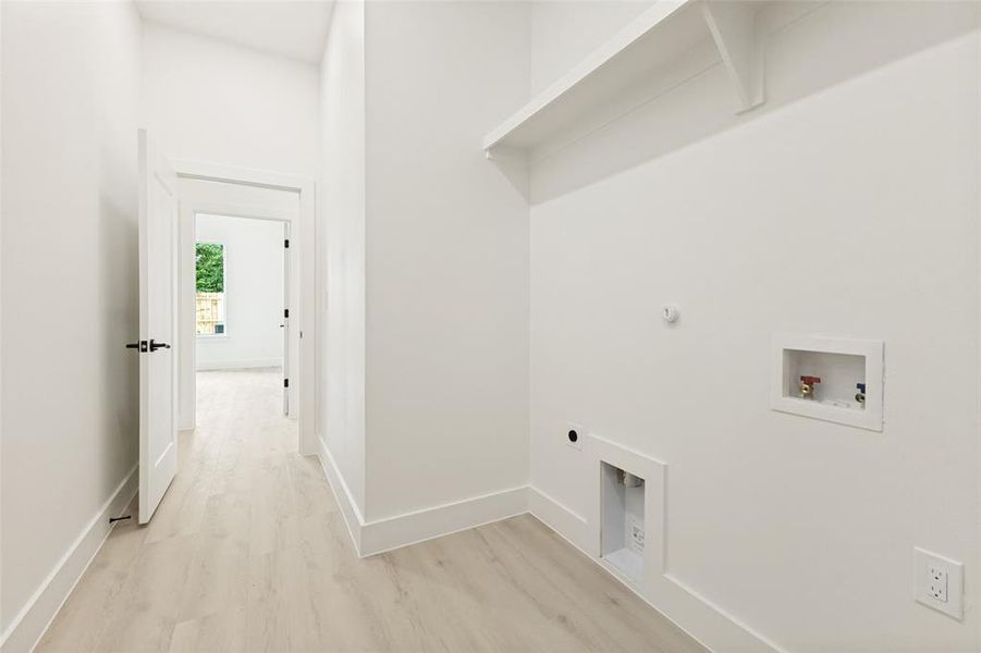Dedicated utility space featuring a built-in shelving unit, light-toned wood-finish flooring, white walls, and a full-size laundry hookup Dedicated utility space featuring a built-in shelving unit, light-toned wood-finish flooring, white walls, and a full-size laundry hookup