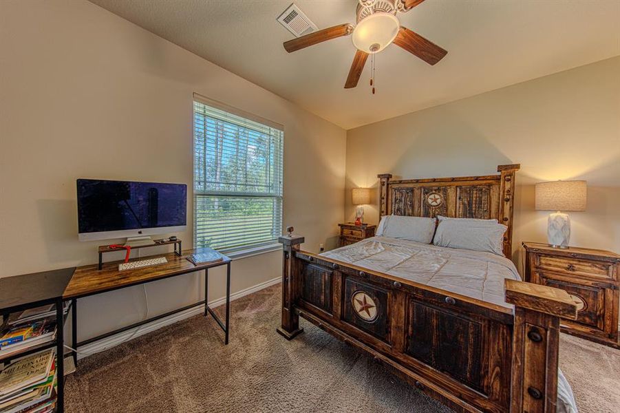 This cozy bedroom features rustic wooden furniture, including a bed and nightstands, complemented by a ceiling fan. A large window allows natural light to brighten the space, which also includes a compact workspace with a desk and computer.