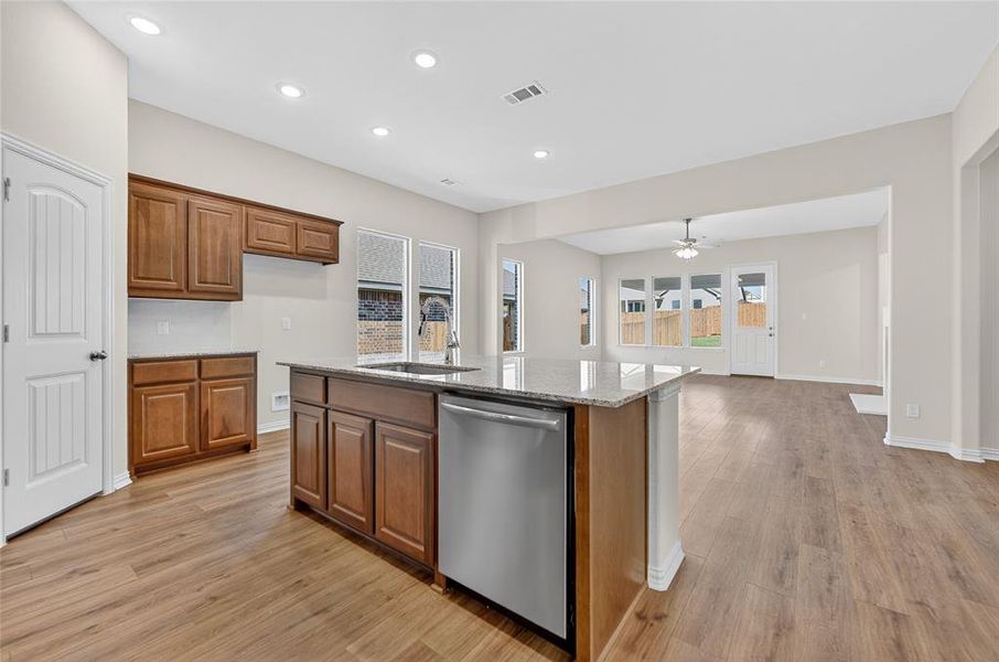 Kitchen with brown cabinetry, light stone countertops, stainless steel dishwasher, open floor plan, and light wood finished floors