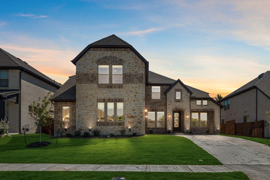 Two-story home with stone and brick exterior, dark roof, arched entry, and manicured front lawn at dusk