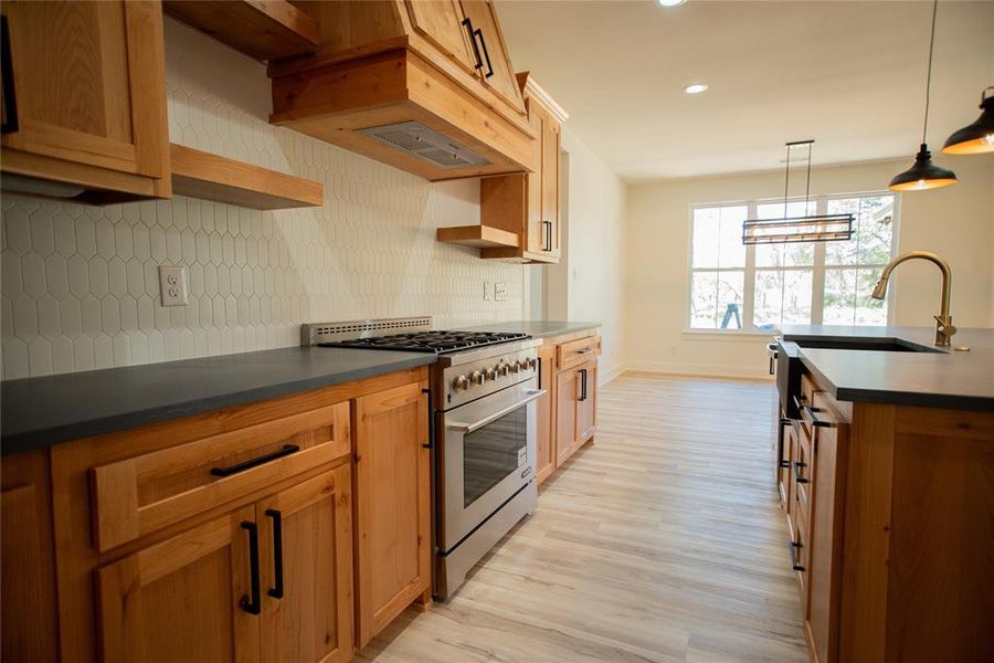 Kitchen featuring tasteful backsplash, light wood-type flooring, high end stove, hanging light fixtures, and dark countertops Kitchen featuring tasteful backsplash, light wood-type flooring, high end stove, hanging light fixtures, and dark countertops