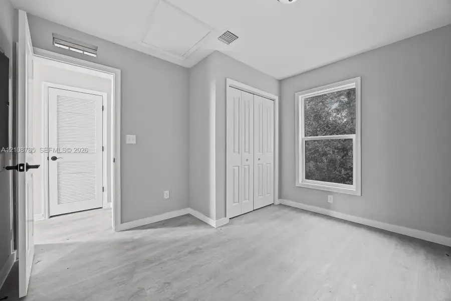 Interior space featuring neutral wall paint, wood-finish flooring, double-panel closet doors, and a single-hung window with exterior greenery Interior space featuring neutral wall paint, wood-finish flooring, double-panel closet doors, and a single-hung window with exterior greenery