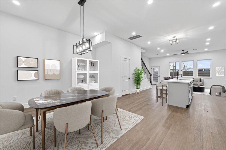 This modern open-concept space features a dining area with elegant lighting, a bright kitchen with a large island and bar seating, and a cozy living area. The room is enhanced by natural light from several windows, creating an inviting atmosphere.