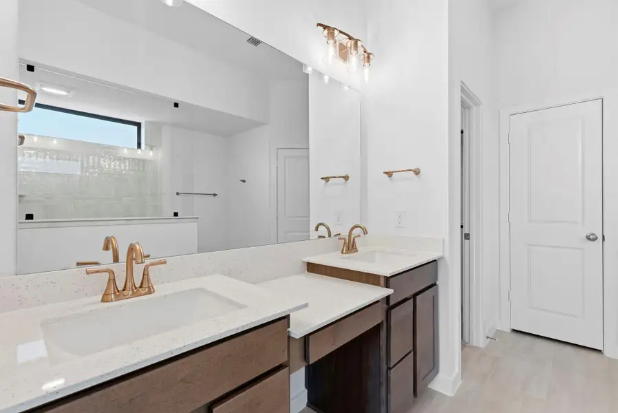 Double sinks with a convenient vanity between. This main bathroom is outfitted with style and elegance featuring beautiful natural wood cabinets, white quartz countertops, and modern gold fixtures throughout.