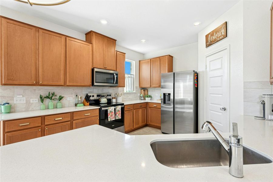 The kitchen highlights sleek stainless steel appliances, warm wood-finish cabinetry, a stylish decorative backsplash, light tile-patterned flooring, and beautiful light stone countertops—creating a bright, inviting, and well-appointed culinary space.