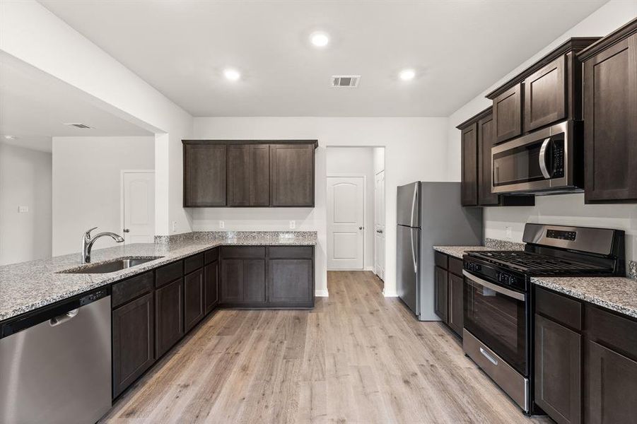 Kitchen featuring stainless steel appliances, dark brown cabinetry, light stone counters, light wood-style flooring, and recessed lighting