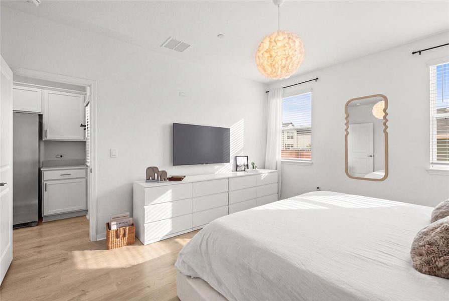Sun-filled bedroom featuring light wood flooring and a luminous pendant light fixture. Some elements digitally staged.