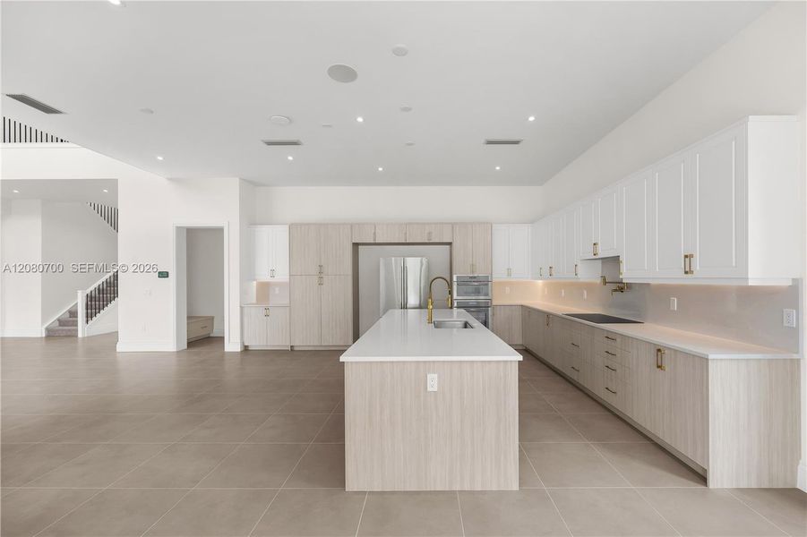 Spacious kitchen featuring an expansive island with a built-in sink, stainless steel appliances, extensive cabinetry with integrated lighting, and neutral-toned floor tiling