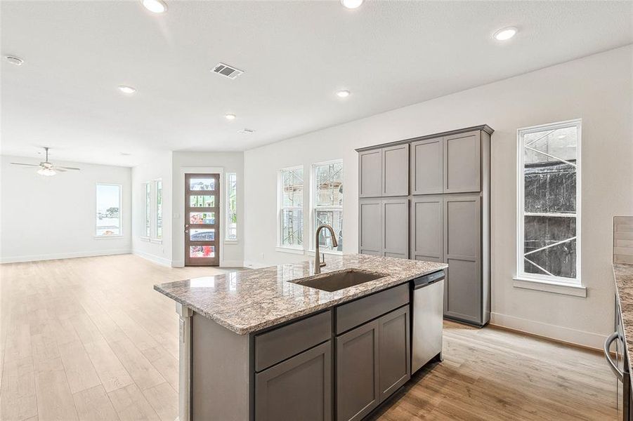 Kitchen with stainless steel dishwasher, sink, light wood-type flooring, light stone countertops, and a kitchen island with sink