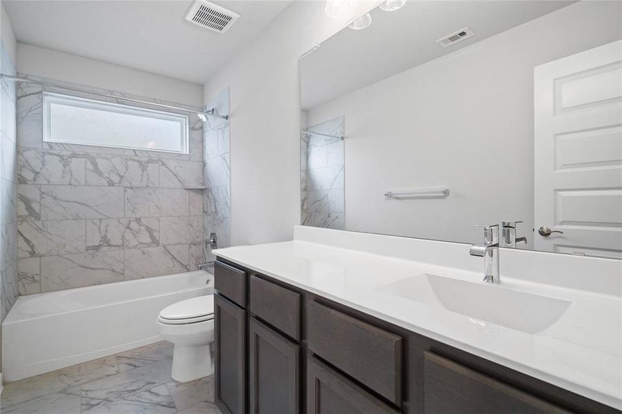 Secondary bath features tile flooring, bath/shower combo with tile surround, stained wood cabinets, beautiful light countertops, mirror, dark, sleek fixtures and modern finishes.