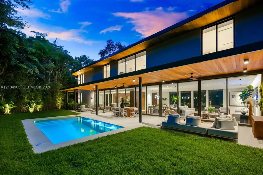 Modern architectural exterior featuring expansive wood-paneled eaves, full-height sliding glass walls, a covered concrete patio, and an in-ground swimming pool set within a manicured lawn