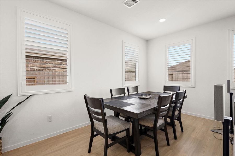 This dining area offers a bright, modern space with light wood flooring, white walls, and three large windows with shutters. It features a sleek dark wood table set for six, creating an inviting atmosphere for meals.