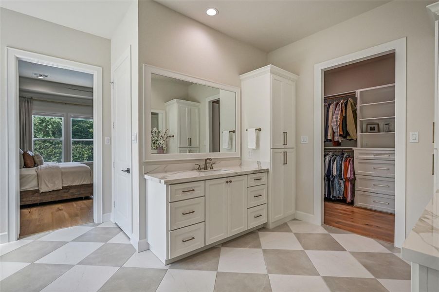 The beautifully appointed primary bath features separate vanities with abundant cabinetry, stone-look countertops and oversized mirrors, plus a striking large-format checkerboard tile floor and direct access to two separate customized closets.