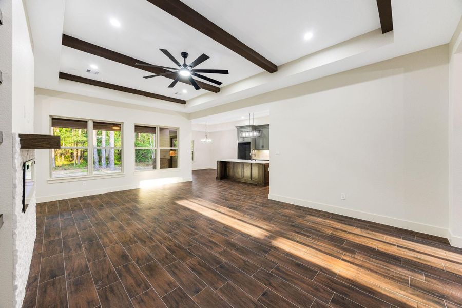 Spacious, open-concept living area with large windows offering natural light and scenic views. Features wood-look tile flooring, a modern ceiling fan, and exposed beams. The kitchen area includes contemporary lighting and ample space for dining.