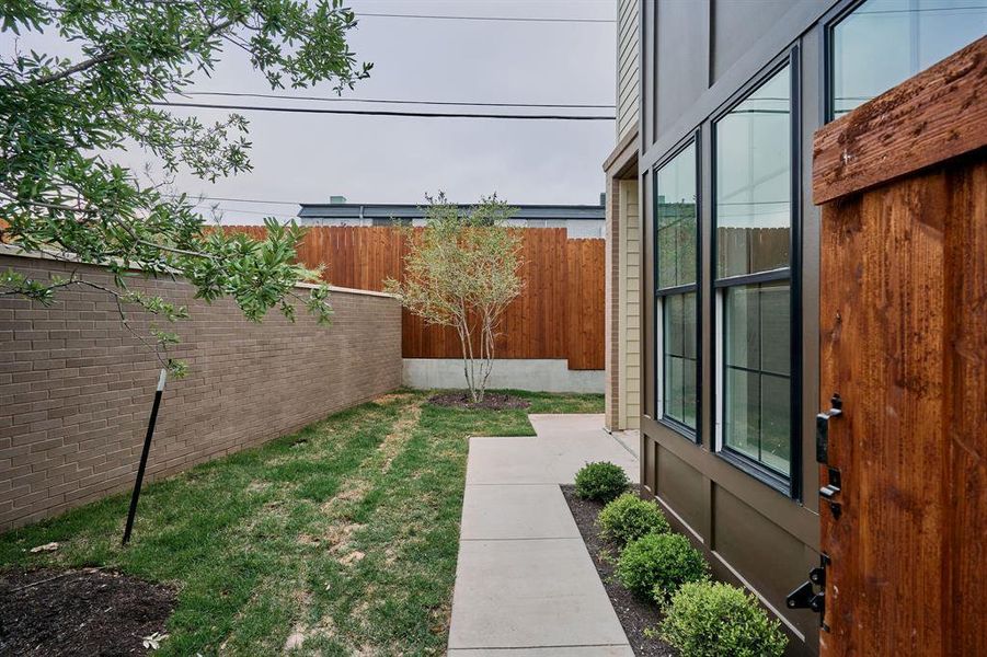Exterior side yard featuring a concrete walkway, established shrubs, a modern wood privacy fence, and a brick retaining wall
