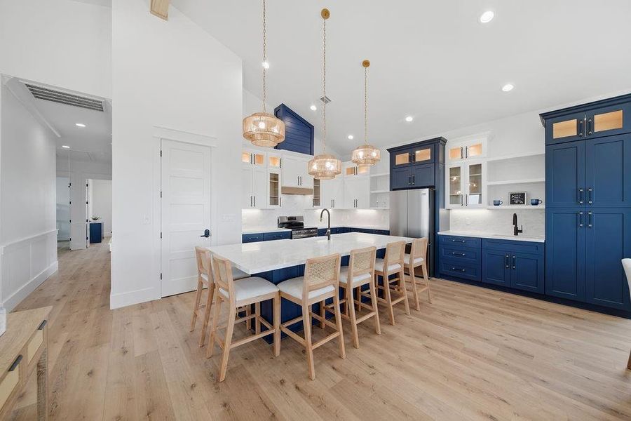 This modern kitchen features sleek blue cabinetry, a spacious white island with seating for six, elegant pendant lighting, and stainless steel appliances. The open layout and light wood flooring enhance its inviting atmosphere.