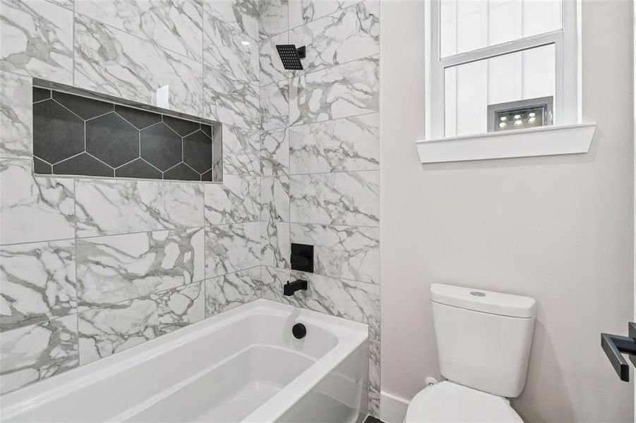 Bathtub with marble-patterned tile surround, recessed shower niche with hexagonal tile, matte black shower fixtures, and a white toilet