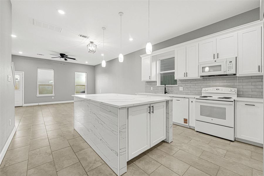 Modern kitchen with sleek white cabinetry, a spacious island with marble-like countertops, and appliances. The open layout flows into a bright living area with tiled floors, recessed lighting, and a ceiling fan, creating a welcoming and functional space.