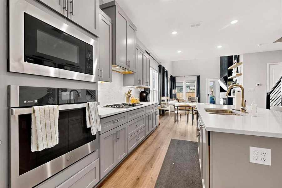 Kitchen featuring stainless steel appliances, gray cabinets, decorative backsplash, light wood-type flooring, and light stone countertops Kitchen featuring stainless steel appliances, gray cabinets, decorative backsplash, light wood-type flooring, and light stone countertops