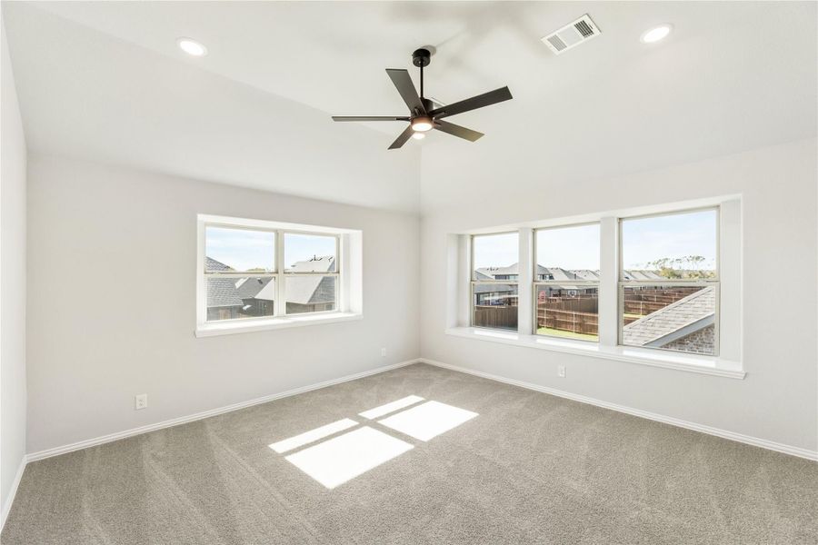 Empty bedroom with vaulted ceiling, ceiling fan, carpet flooring, and multiple windows with neighborhood views