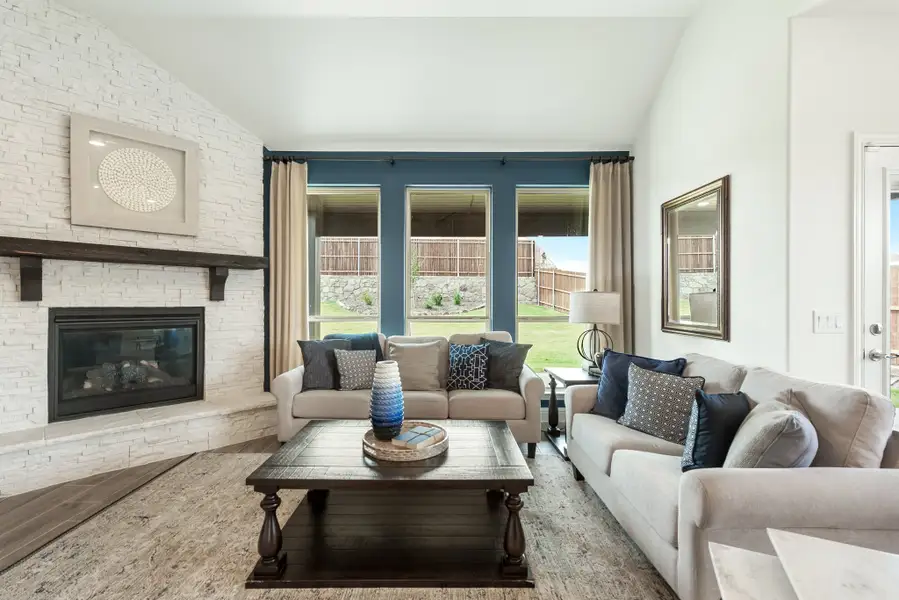 Living room with vaulted ceiling, white stone fireplace, two beige sofas, and large windows with blue accent wall