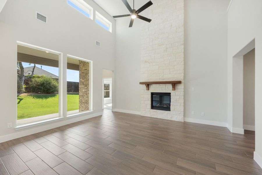 Open living room with two-story ceiling, stone fireplace, ceiling fan, and large windows overlooking backyard Open living room with two-story ceiling, stone fireplace, ceiling fan, and large windows overlooking backyard
