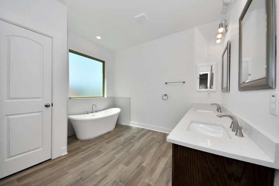 Spacious, modern bathroom featuring a freestanding tub, dual sinks with sleek fixtures, and elegant ceramic wood-look flooring. Bright and inviting with frosted window for privacy.
