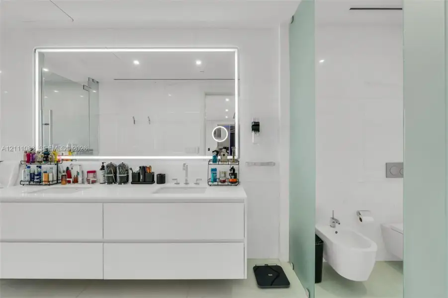 Floating white vanity with dual rectangular undermount sinks, large LED-backlit mirror, wall-to-wall stone-finish tiling, and wall-mounted bidet and toilet Floating white vanity with dual rectangular undermount sinks, large LED-backlit mirror, wall-to-wall stone-finish tiling, and wall-mounted bidet and toilet