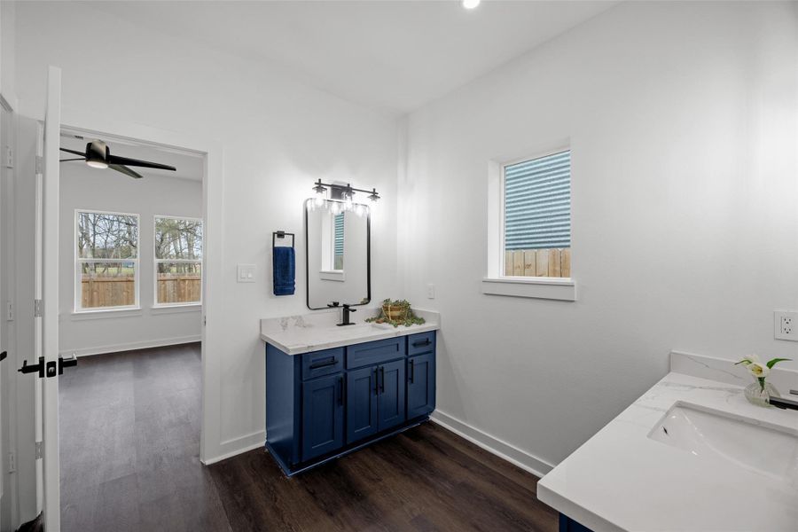 Vibrant white walls and dark wood flooring. It features a stylish blue vanity with dual sinks and a large mirror. The room is connected to another space via a door, offering natural light from two windows. Vibrant white walls and dark wood flooring. It features a stylish blue vanity with dual sinks and a large mirror. The room is connected to another space via a door, offering natural light from two windows.