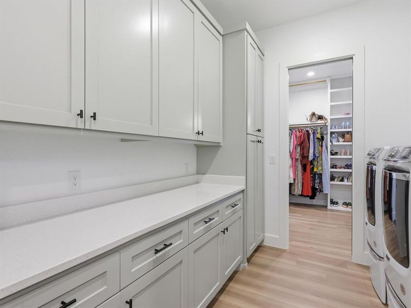 Laundry area featuring extensive white shaker cabinetry, light-toned countertops, and wood-finish flooring, access to primary closet