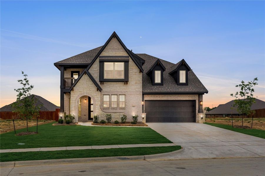 Two-story stone and brick home with dark trim, arched entry, two-car garage, and manicured lawn at dusk Two-story stone and brick home with dark trim, arched entry, two-car garage, and manicured lawn at dusk