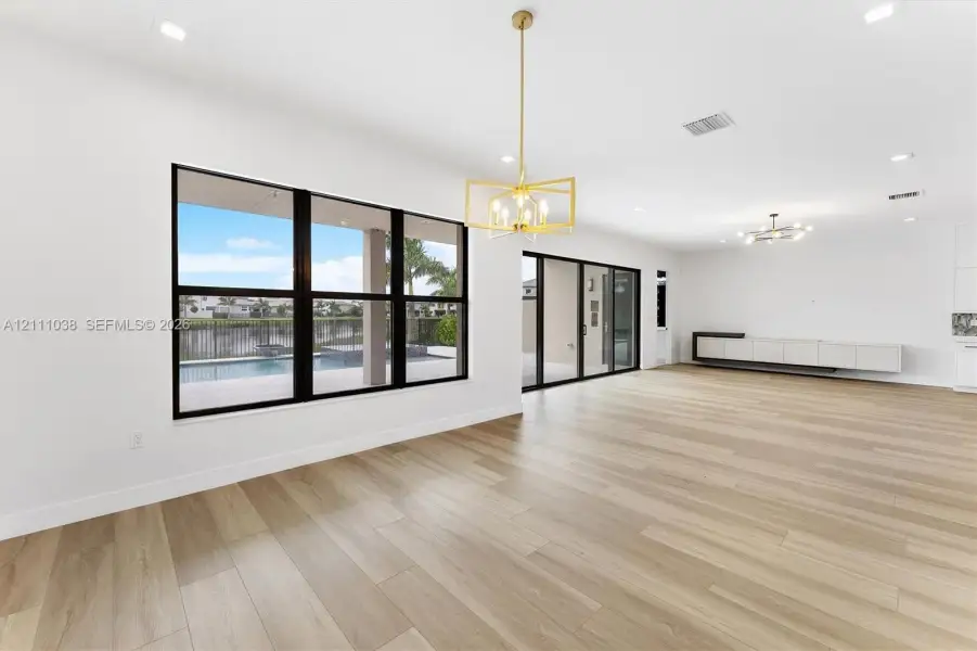 Open-concept interior featuring light wood-finish flooring, large multi-pane windows with dark frames, a geometric metallic pendant light, recessed ceiling lighting, and a built-in wall-mounted cabinet