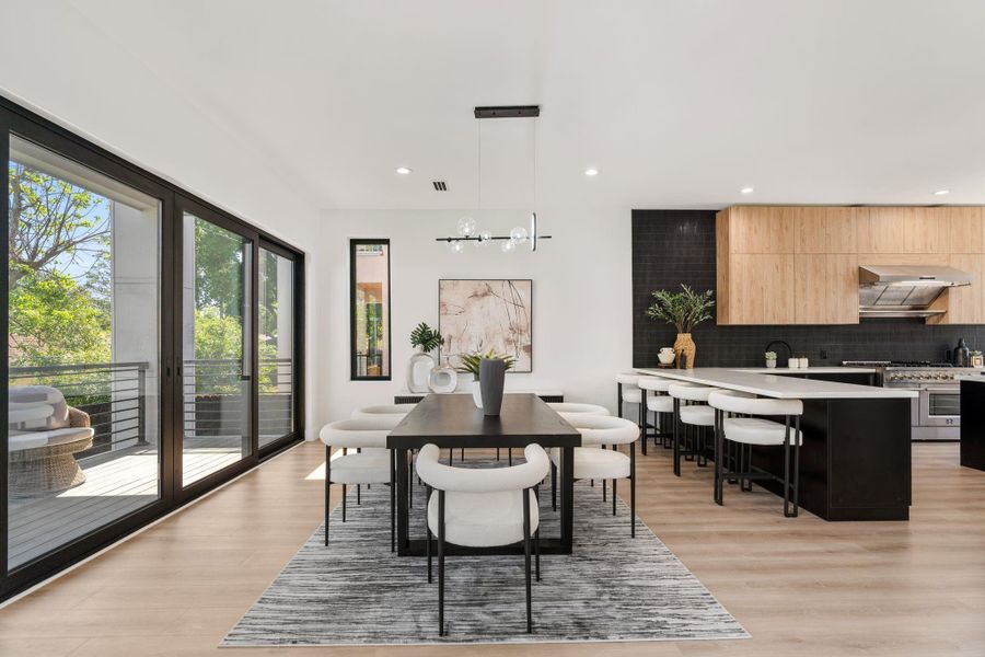 The dining room sits at the natural center of the home's first floor — a modern multi-arm pendant chandelier overhead, the kitchen's warm wood upper cabinets and dark cabinetry framing one side, and a sliding glass door to the covered balcony opening directly on the other. It is a space everything flows through.