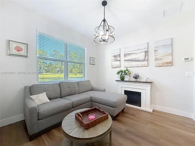 Wood-finish flooring, white fireplace surround with integrated mantel, large window with horizontal blinds, and a multi-light metal pendant fixture