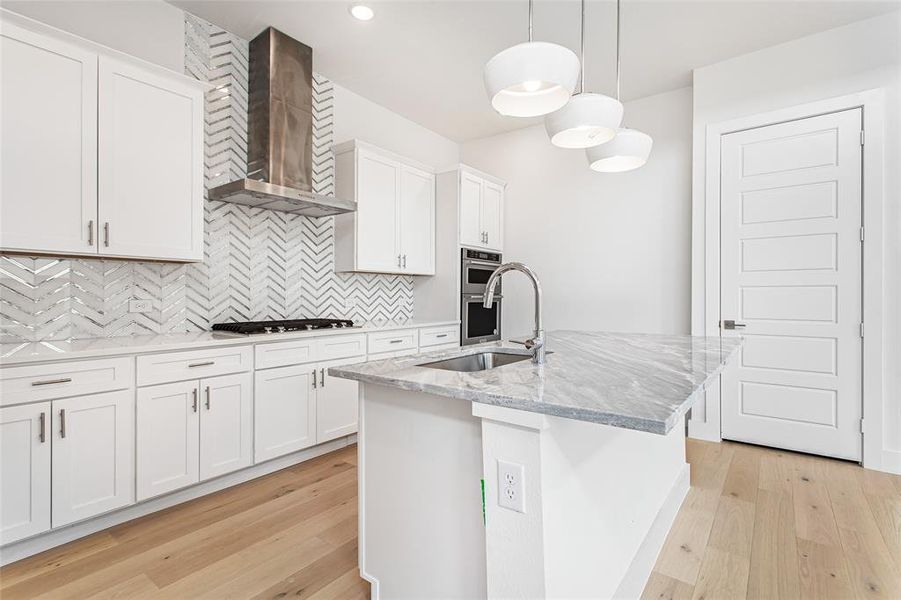 Kitchen featuring light wood-type flooring, a center island with sink, backsplash, white cabinets, and wall chimney range hood