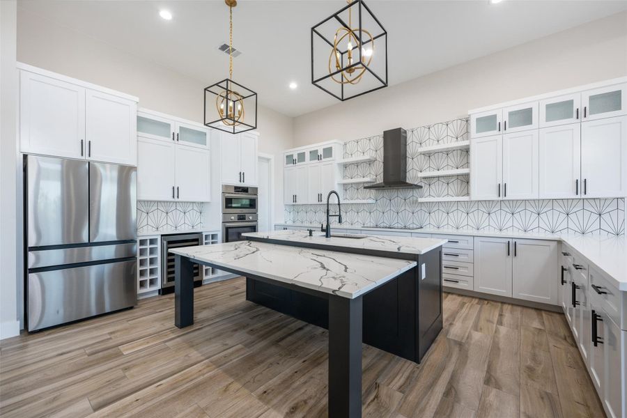Kitchen featuring appliances with stainless steel finishes, hanging light fixtures, white cabinets, a center island with sink, and wall chimney exhaust hood