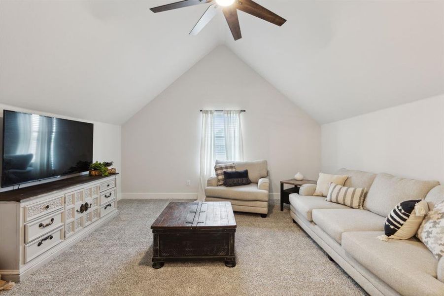 Spacious room featuring vaulted ceilings, light-toned carpeting, a window with horizontal blinds and white drapes, and a ceiling fan with integrated lighting