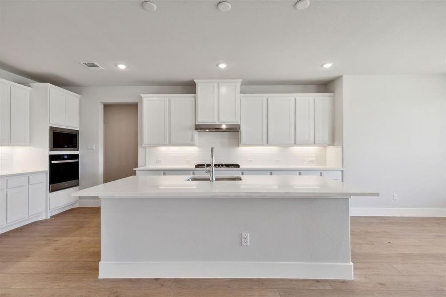 Kitchen with white cabinetry, stainless steel microwave, backsplash, an island with sink, and light hardwood / wood-style floors