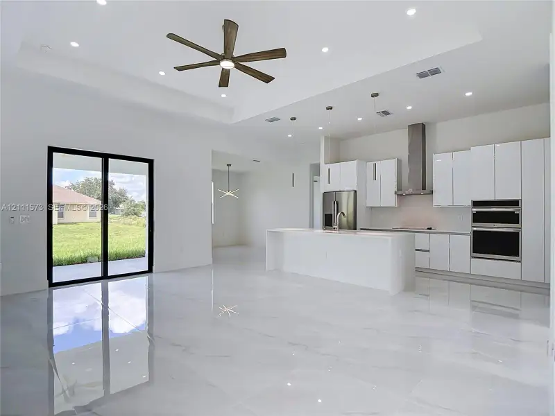 Open-concept living space featuring high tray ceilings, large-format polished tile flooring, white cabinetry, a center island with stone-finish countertops, and built-in stainless steel appliances