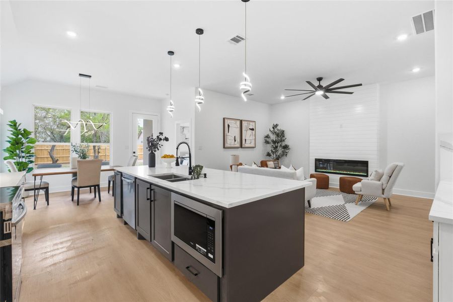 Kitchen featuring light stone countertops, an island with sink, stainless steel appliances, decorative light fixtures, and light wood-style flooring